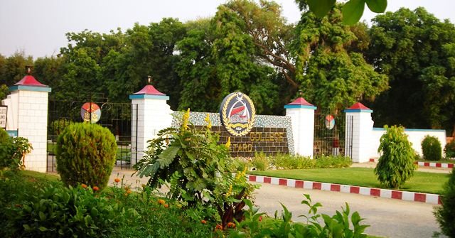 The gate of Cadet College Petaro with the college crest mounted above the mosaic wall, framed by gardens.